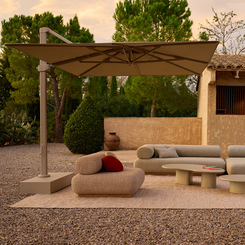 A serene Mediterranean-style outdoor lounge area featuring comfortable wicker furniture, an oversized umbrella, and lush greenery, creating a cozy and inviting atmosphere for outdoor relaxation.
