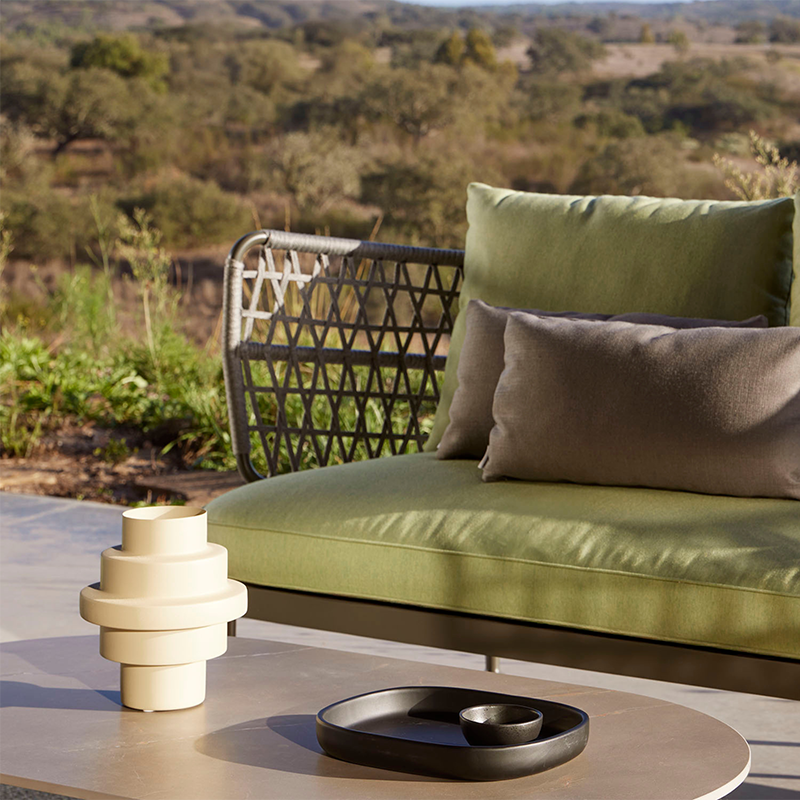 Elegant outdoor lounge furniture featuring a woven metal chair and sofa with plush olive green and neutral cushions, set against a serene natural landscape for a relaxing and inviting ambiance. The contemporary accessories, including a sculptural side table and decorative items, complement the modern outdoor living space.