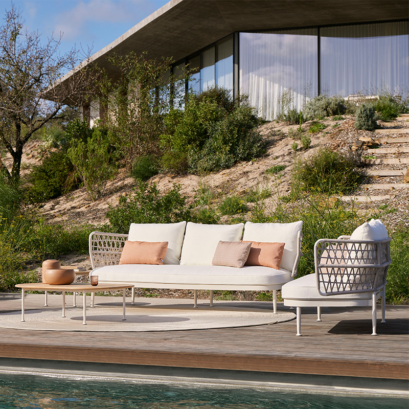 Luxurious outdoor lounge area featuring a stylish modular sofa and armchair set with a woven metal frame and plush, neutral cushions, complemented by a wooden deck and surrounded by a lush, natural landscape for a serene and relaxing atmosphere.