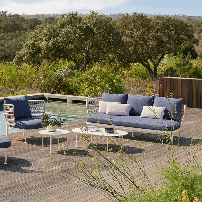 Elegant outdoor lounge space featuring a stylish woven metal patio sofa and armchair set with plush navy blue cushions, surrounded by lush greenery and a serene natural landscape, creating a tranquil and inviting ambiance for relaxation.