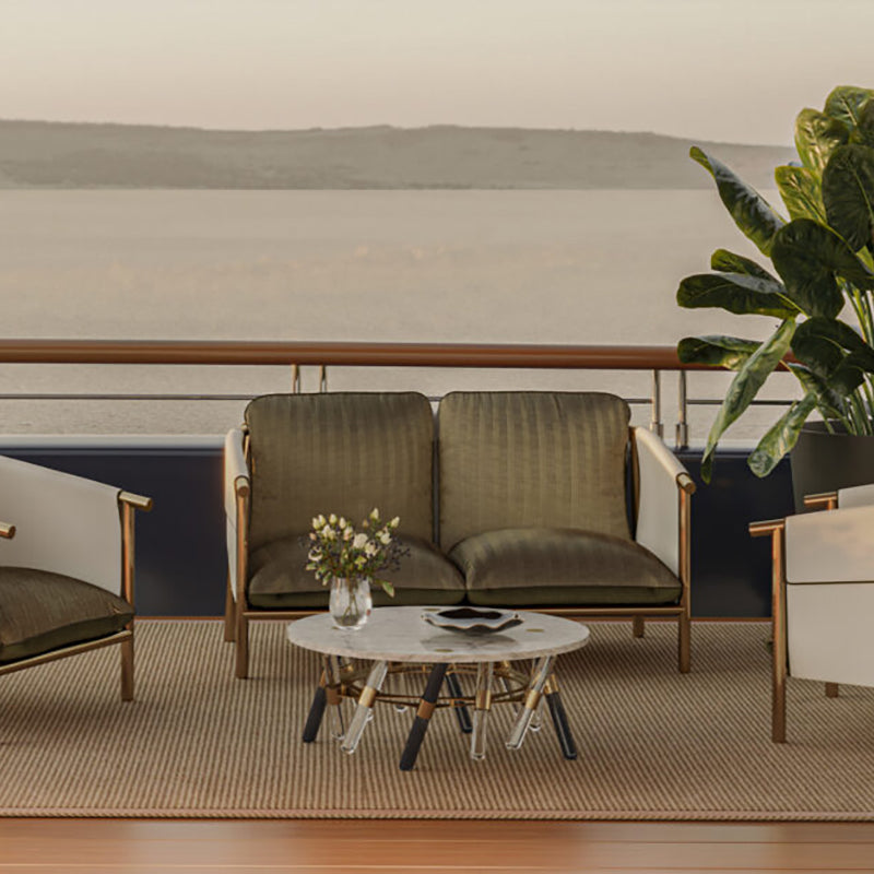 The image depicts a cozy outdoor seating area with a breathtaking view of the landscape in the background. The focal point is a pair of comfortable green armchairs placed on a patterned rug, with a low marble-topped coffee table in front. A small vase of flowers adds a touch of nature to the scene. The setting is framed by a wooden railing, which suggests this is a balcony or deck overlooking the distant hills and water. The overall atmosphere is one of relaxation and appreciation for the natural surroundin