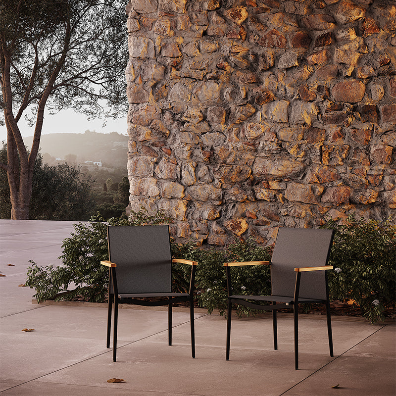 Two sleek, modern outdoor chairs placed in front of an imposing stone wall, with lush greenery and a scenic background visible, creating a serene and stylish outdoor setting.