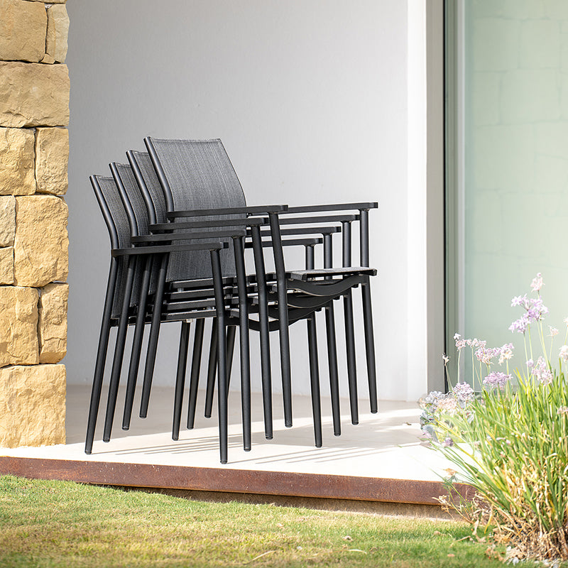 Stacked black metal and mesh chairs arranged on a paved outdoor area, with a stone wall and lush greenery in the background, creating a modern and minimalist design aesthetic.