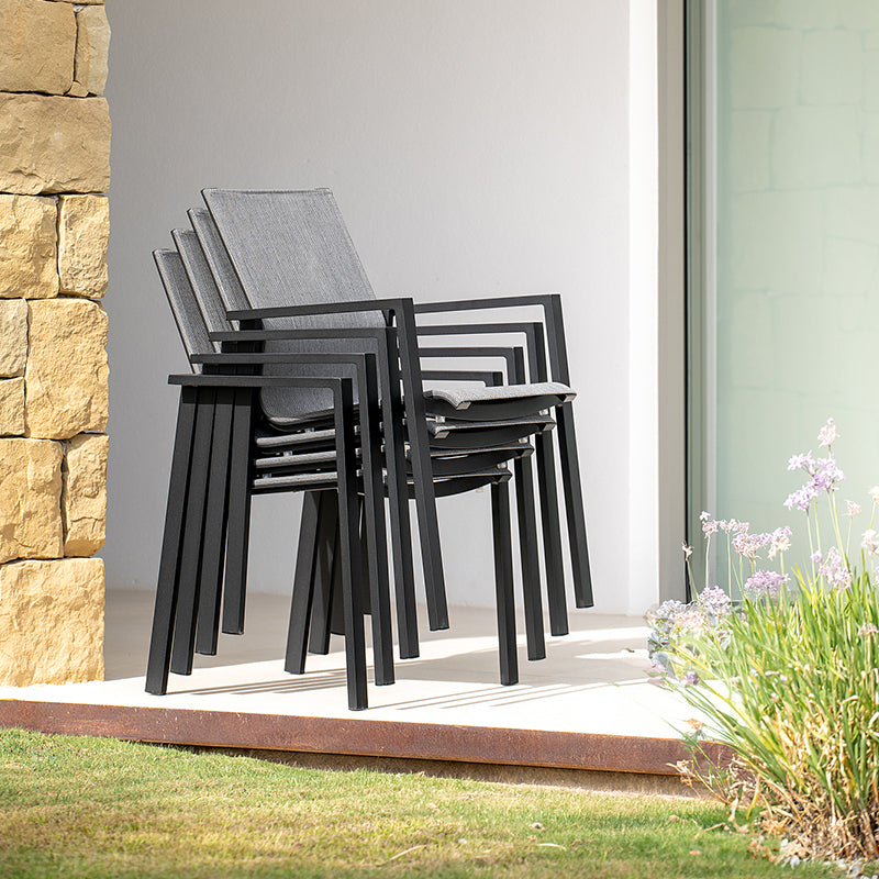 Stacked black metal chairs with gray woven seats, arranged on a stone patio surrounded by greenery.