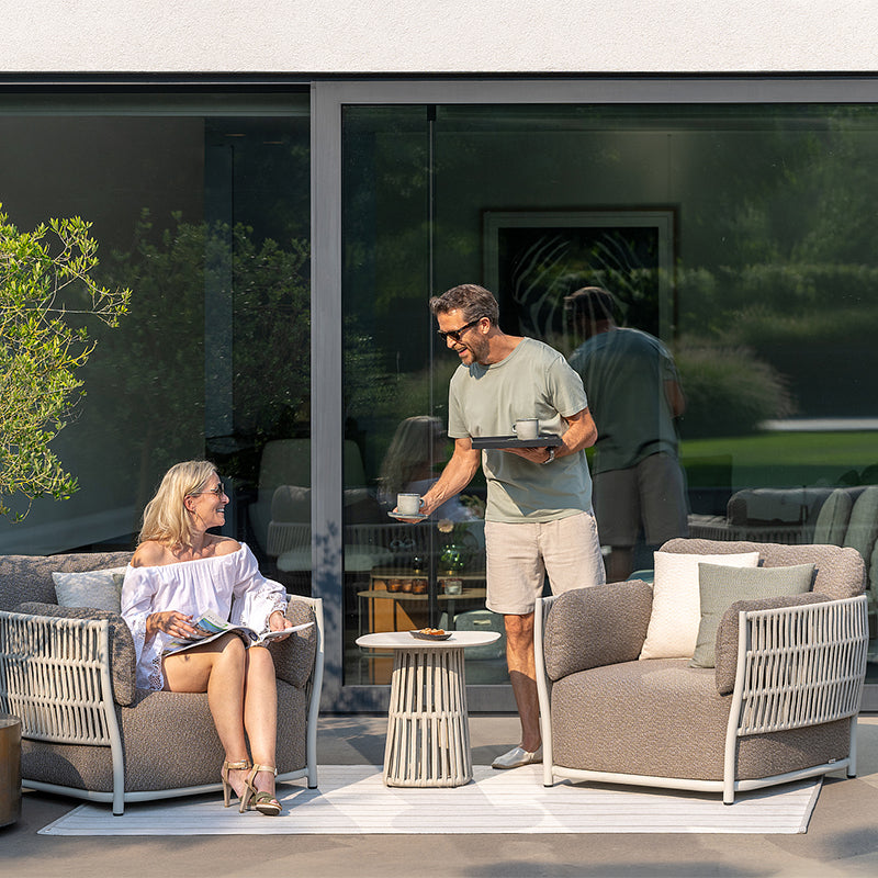 The image depicts a relaxing outdoor living space with a mature couple enjoying each other's company. The scene features contemporary, woven patio furniture set against a lush, natural backdrop, creating an inviting and serene atmosphere for the couple's leisurely afternoon.