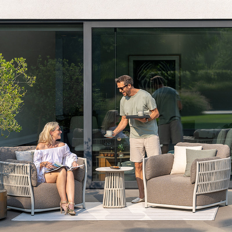 A man in casual clothing serving coffee to a woman relaxing on an outdoor patio with comfortable patio furniture in the foreground, surrounded by lush greenery visible through the glass windows in the background.