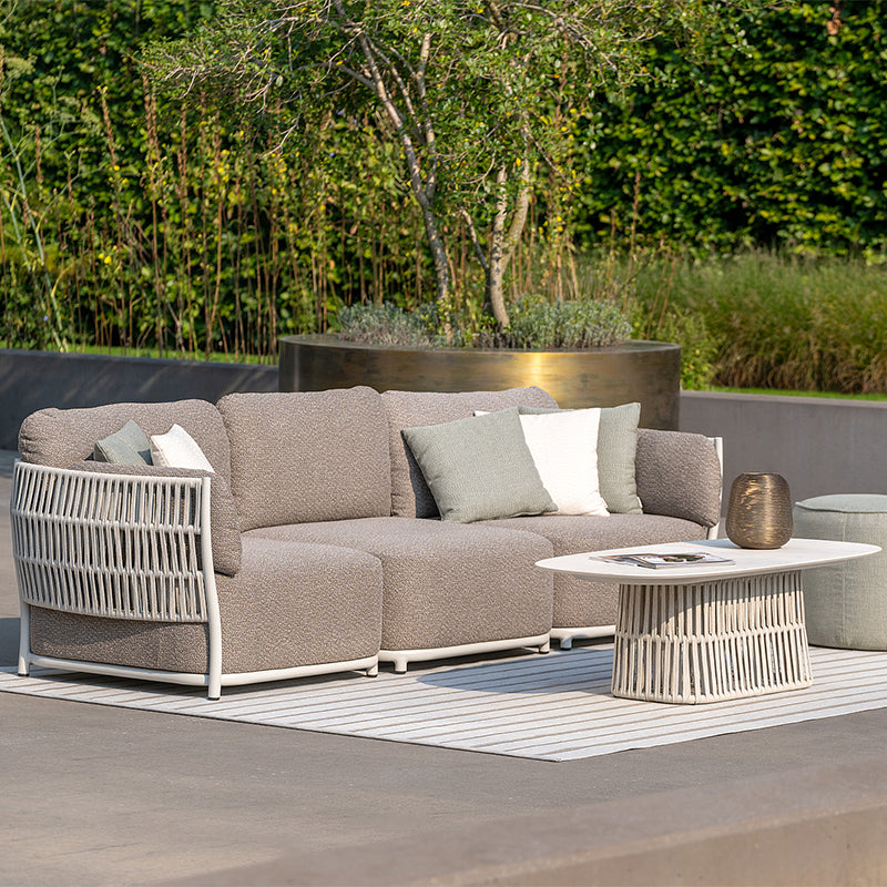Elegant outdoor lounge furniture set with beige upholstered sofa and matching armchair, placed on a paved patio against a lush, natural backdrop.