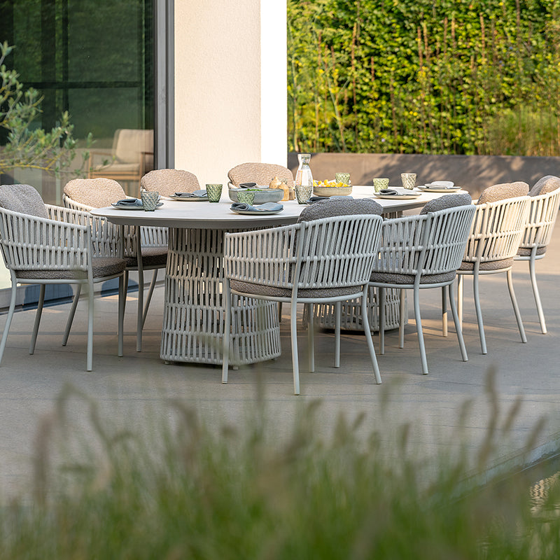 This image showcases an elegant Outdoor Furniture dining set, featuring a large oval table surrounded by stylish woven chairs with plush gray cushions. The natural setting, with lush greenery and a glass wall, creates a serene and inviting atmosphere for outdoor dining and entertaining.