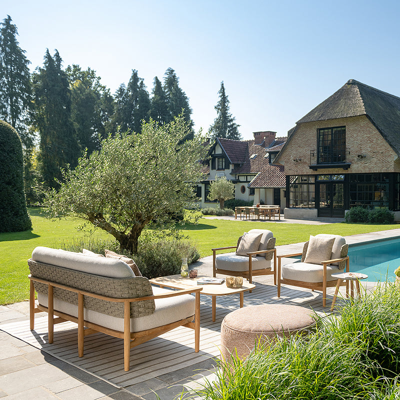 Serene outdoor living space with comfortable wooden furniture and plush cushions surrounding a swimming pool, framed by lush greenery and a charming rustic architectural structure in the background.