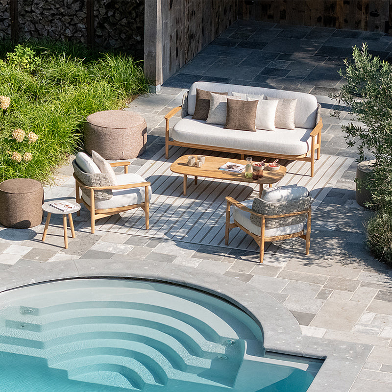 Luxurious outdoor lounge area with comfortable wooden furniture, plush cushions, and a decorative pool with a unique ripple design, all set against a backdrop of lush greenery and stone walls.