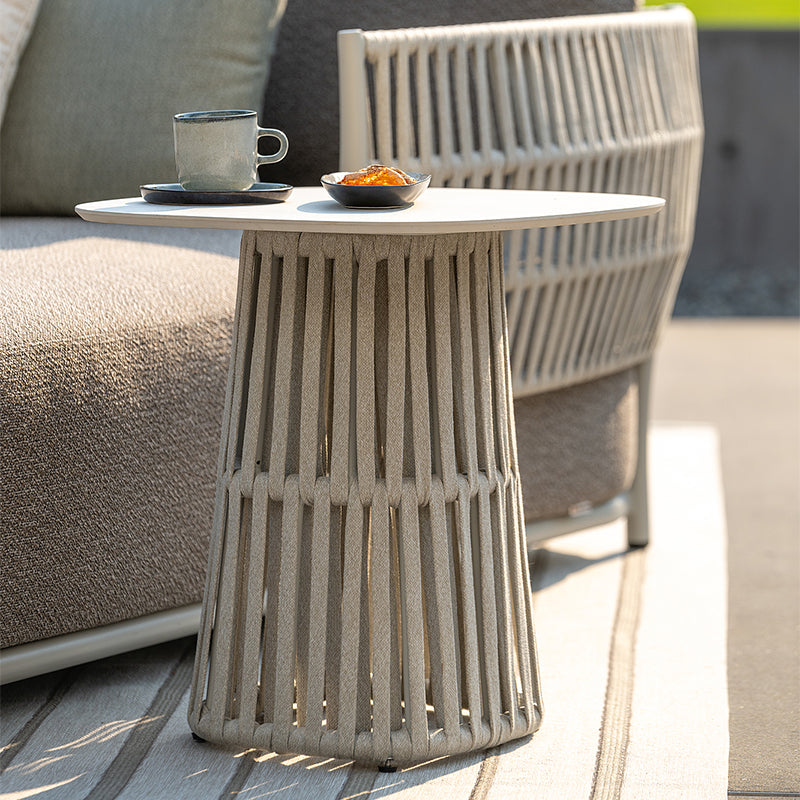 A modern, minimalist outdoor side table with a ribbed concrete base and a round white tabletop, displaying a coffee cup and a small dish on it.