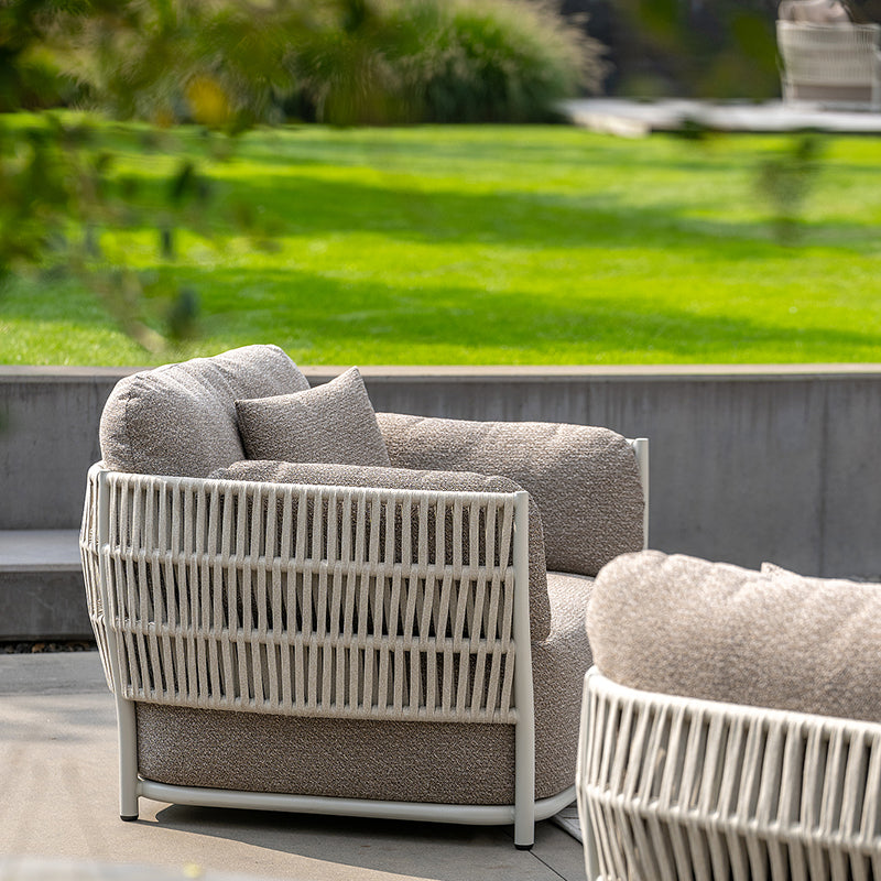 This image shows a pair of modern, minimalist outdoor armchairs with clean, slatted frames and plush, cushioned seats. The chairs are positioned on a patio or deck, overlooking a lush, green outdoor space. The design is visually striking and creates a serene, inviting atmosphere.