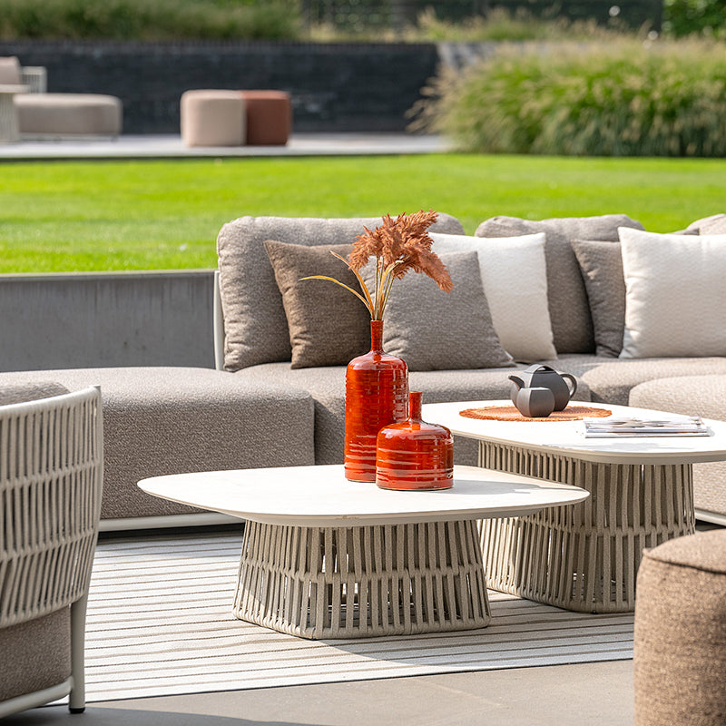 Stylish outdoor patio furniture set with gray cushioned sofas, modern white coffee tables, and decorative accents in a landscaped backyard setting.