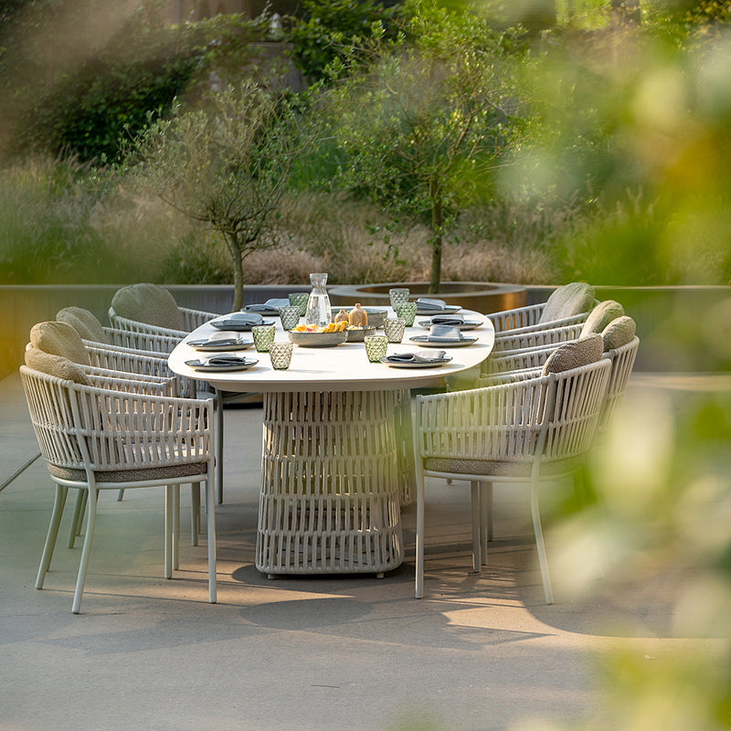 This image depicts an elegant Outdoor Furniture dining set in a lush, natural setting. The oval table and woven chairs create a stylish and inviting al fresco dining experience, surrounded by verdant greenery and a glass wall that allows the natural environment to seamlessly integrate with the living space.