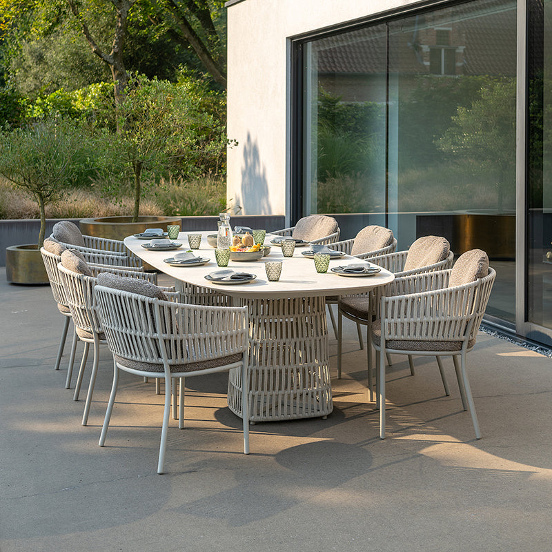 This image showcases a luxurious Outdoor Furniture dining set, featuring a large oval table surrounded by stylish woven chairs with plush cushions. The setting is enhanced by the expansive glass wall that seamlessly integrates the interior and exterior spaces, creating a serene and inviting al fresco dining experience.