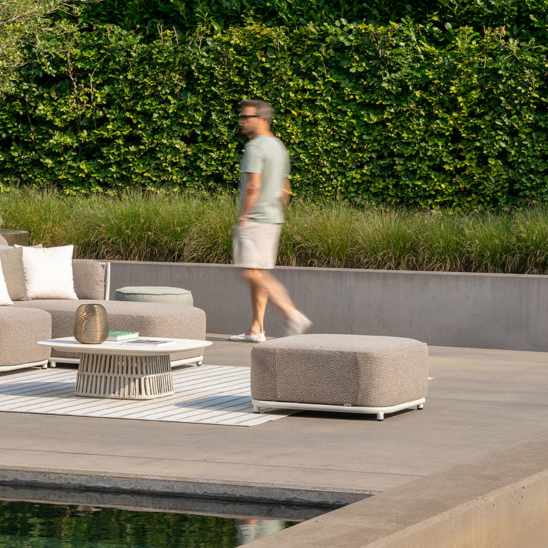An outdoor seating area with modern, woven furniture on a wooden deck surrounded by lush greenery. A person is shown walking past, creating a sense of movement and life within the serene landscape.