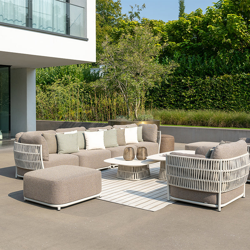 An elegant, contemporary outdoor seating arrangement featuring a modular sofa, armchair, and coffee table. The neutral-toned, woven furniture is set against a backdrop of lush greenery, creating a serene and inviting space.