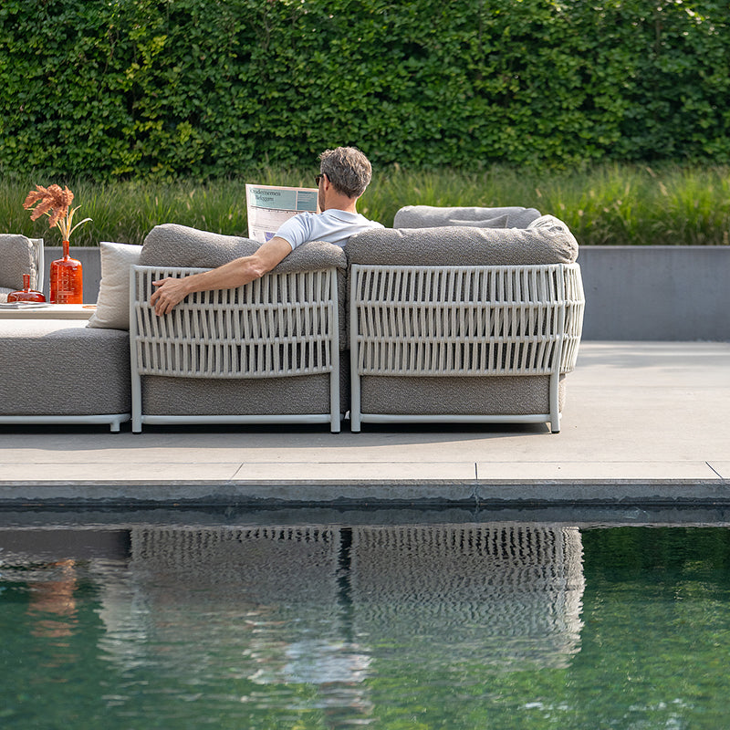 A man relaxing on a modern outdoor sectional sofa, surrounded by a lush green landscape and reflected in the still waters of a pool. The furniture features a minimalist, woven design.