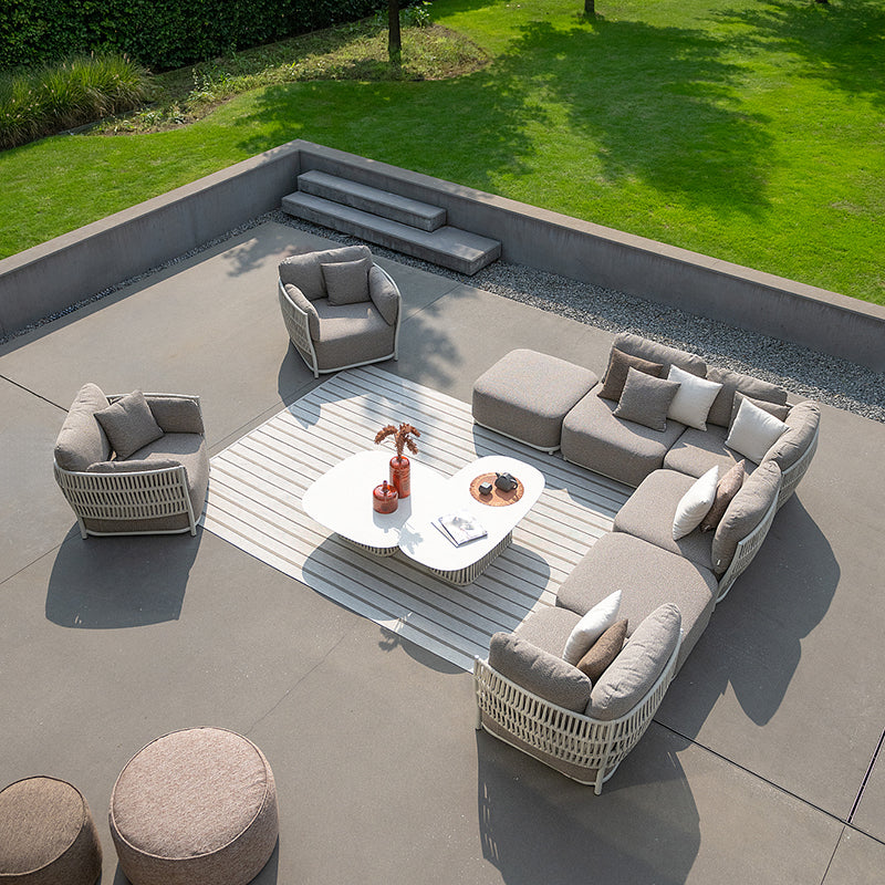 Elegant outdoor living space with a modular sofa set, armchair, and coffee table arranged around a sunken seating area. The modern furniture features a woven design and neutral tones, complementing the lush green landscape surrounding the patio.
