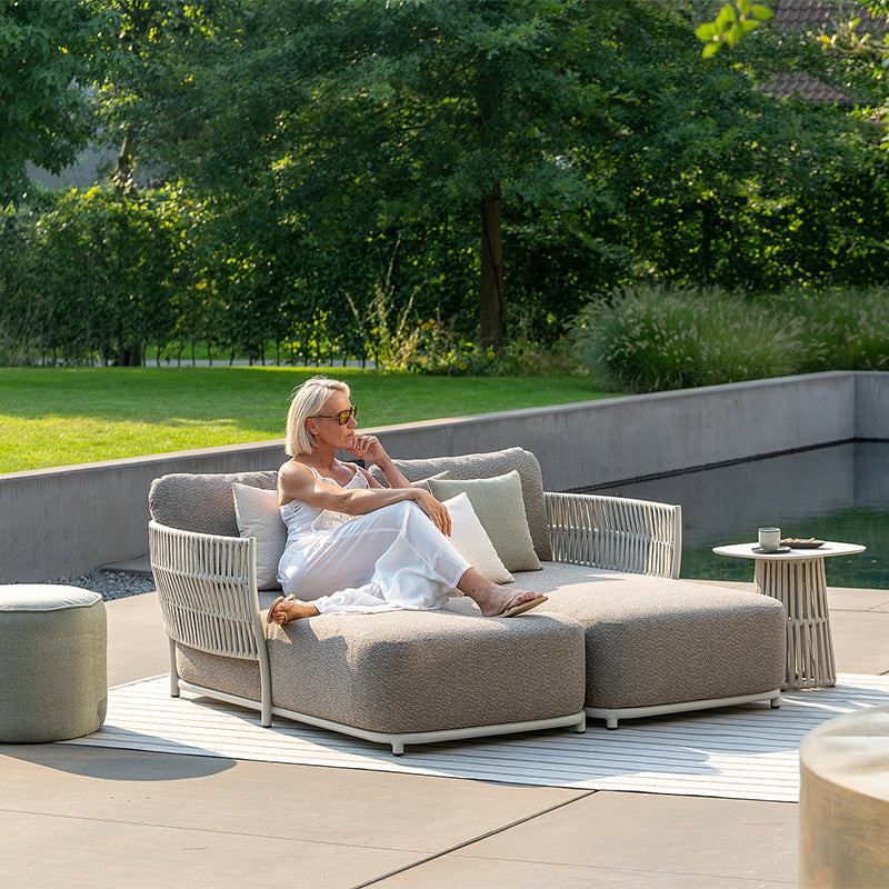 A woman reclining on a comfortable outdoor couch in a lush, green garden setting, surrounded by trees and nature. She appears relaxed and at peace in the tranquil outdoor environment.