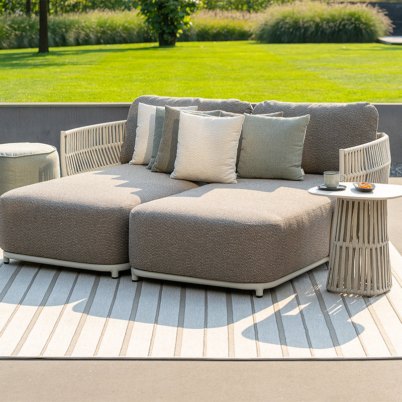 Outdoor patio furniture set with a comfortable, L-shaped gray couch and side tables, situated on a wooden deck overlooking a lush green lawn and garden.