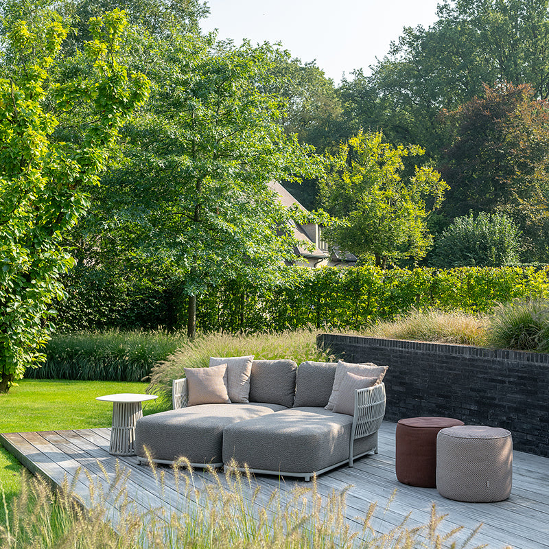 Outdoor patio seating arrangement with a gray modular sofa, side tables, and poufs set on a wooden deck surrounded by lush greenery and a manicured lawn.