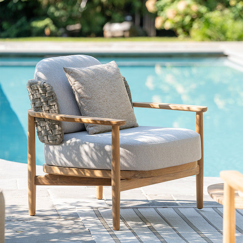 A comfortable, wooden outdoor chair with plush gray cushions placed near a sparkling pool, creating an inviting and relaxing ambiance.
