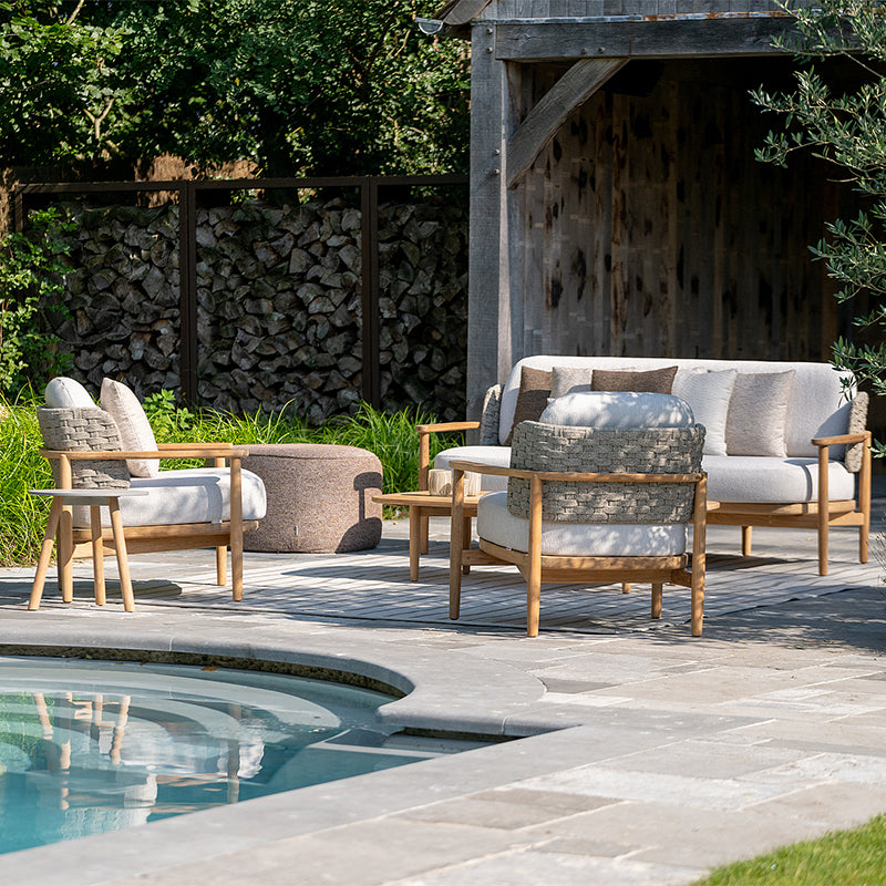  Inviting outdoor lounge area with comfortable wooden furniture and plush cushions, positioned around a decorative pool and surrounded by a stone wall and lush greenery, creating a peaceful and relaxing atmosphere.