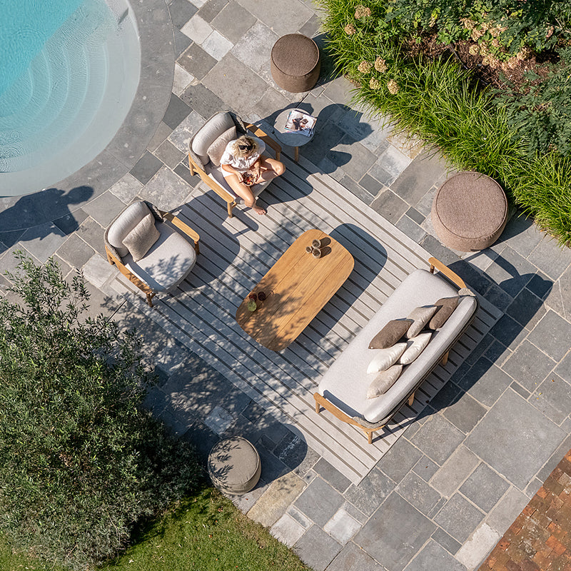 Overhead view of a serene outdoor lounge area with comfortable wooden furniture, decorative poufs, and a sleek wooden coffee table, set against a backdrop of lush greenery and a sparkling swimming pool.