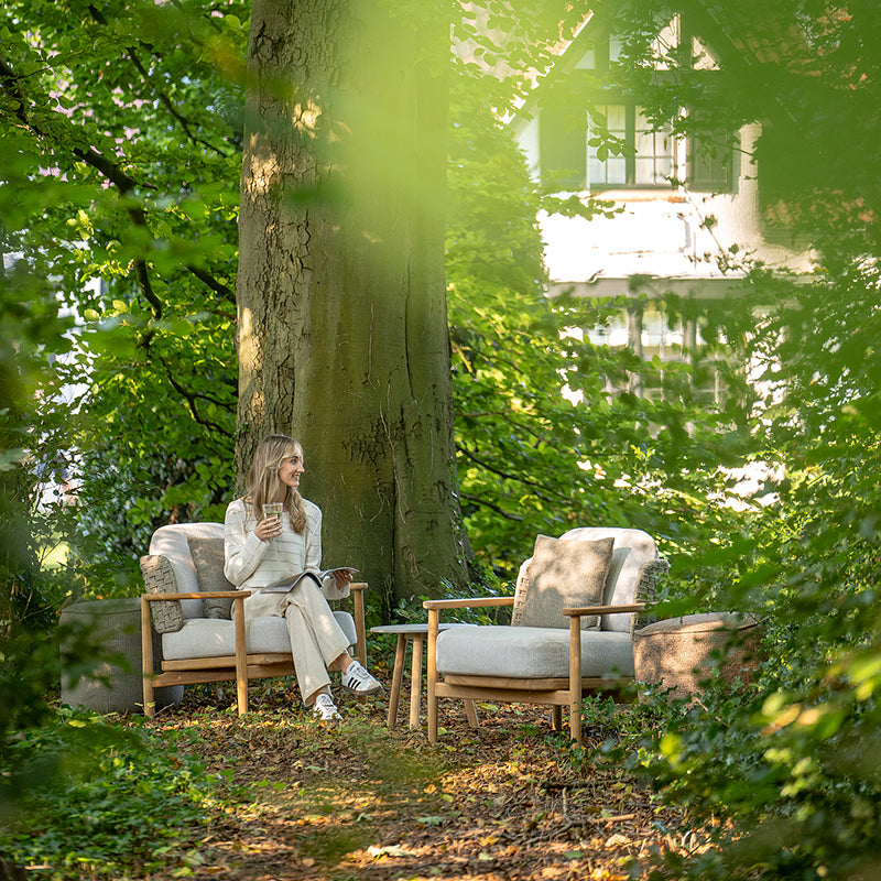 A woman sitting alone on a comfortable outdoor chair, enjoying a beverage amidst the serene natural setting of a lush, wooded area.