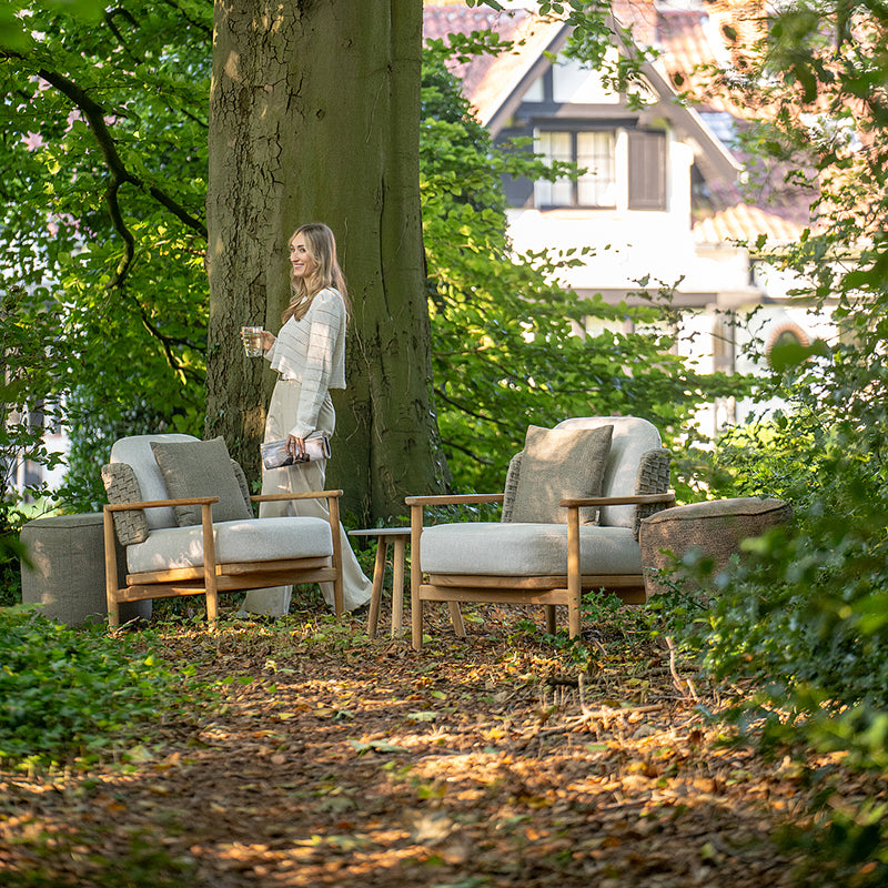 Serene outdoor seating area with comfortable wooden chairs and a person standing near a large tree, surrounded by lush greenery and a charming rustic building in the background, creating a tranquil and inviting atmosphere.