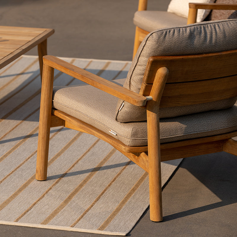 A cozy and stylish outdoor armchair with a natural wood frame and beige upholstered cushions, complemented by a wooden dining table, creating an inviting and harmonious outdoor living space.