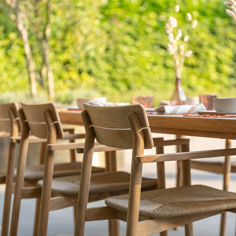 Outdoor dining setup with a wooden table and chairs, surrounded by lush greenery, creating a serene and inviting atmosphere for alfresco dining. The furniture features a natural, minimalist design that blends well with the natural environment.