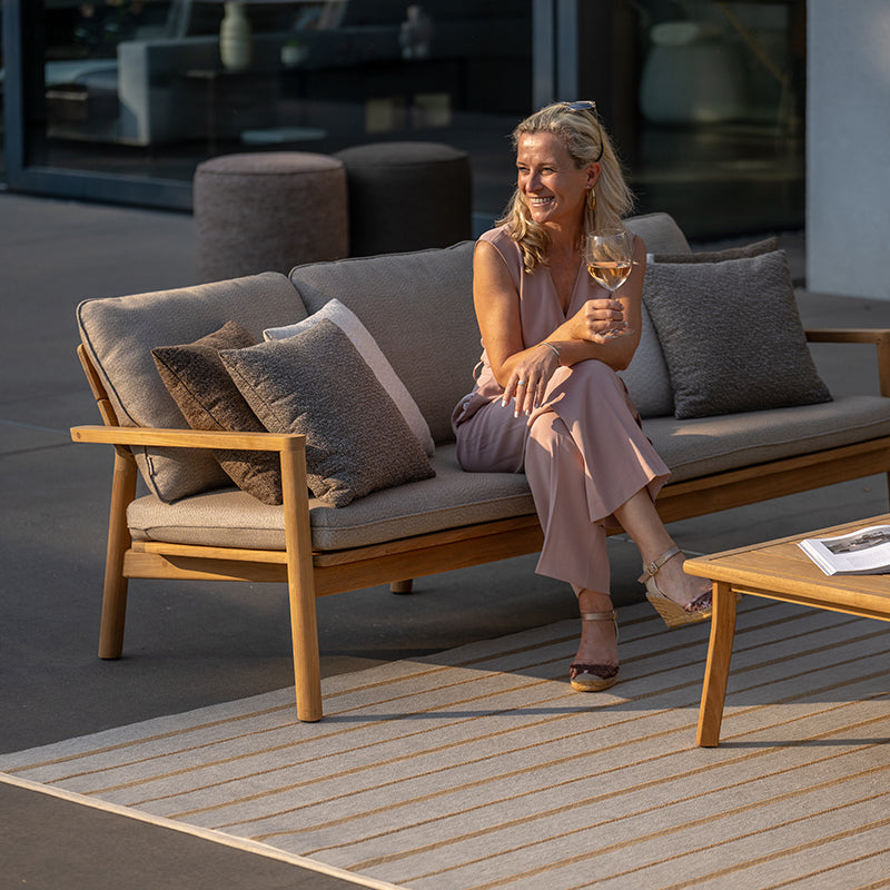 This image showcases a relaxed and inviting outdoor seating arrangement, with a comfortable sofa featuring a natural wood frame and plush, neutral-toned cushions. A smiling woman is seated on the sofa, enjoying a glass of wine and taking in the pleasant surroundings, creating an atmosphere of tranquility and leisure in this Outdoor Furniture setting.
