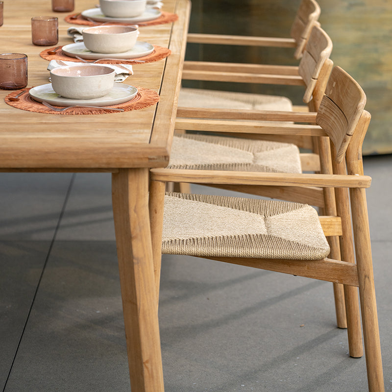 Wooden outdoor dining furniture showcasing a rectangular table with a natural wood finish, accompanied by matching chairs with woven rattan seats. The minimalist design and earthy tones create a warm and inviting atmosphere for alfresco dining, blending seamlessly with the surrounding environment.