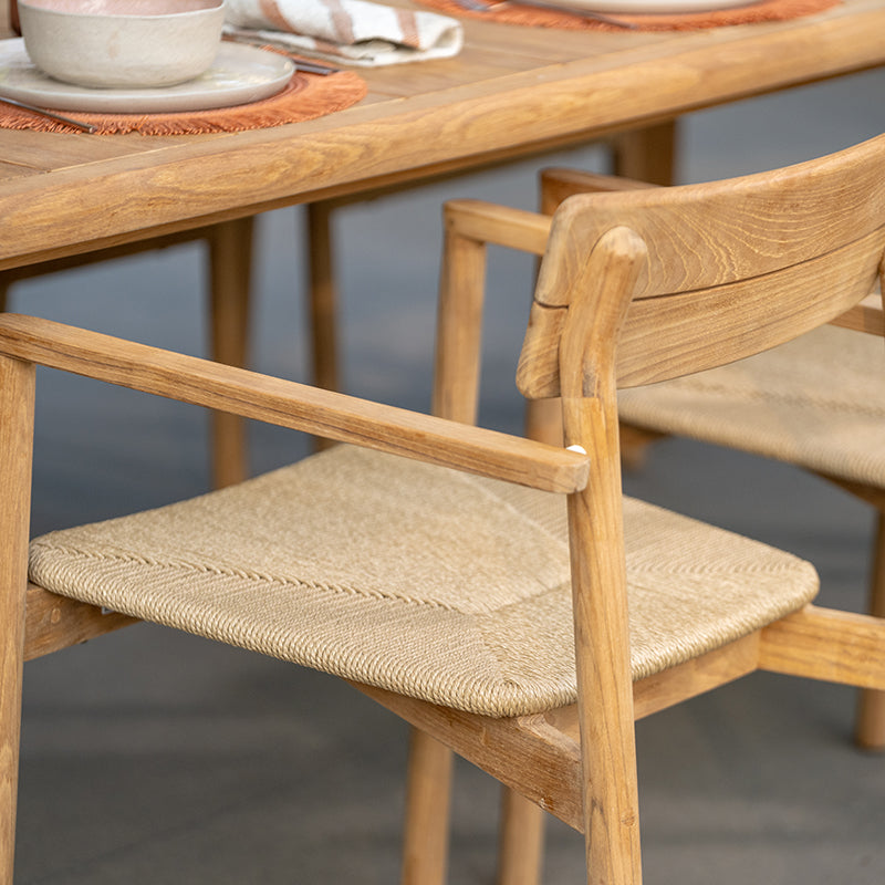 Close-up view of a wooden outdoor dining chair with a woven rattan seat, showcasing the intricate craftsmanship and natural materials used in its construction. The chair's simple, elegant design complements the wooden table and surrounding outdoor setting, creating a cohesive and visually appealing outdoor furniture arrangement.