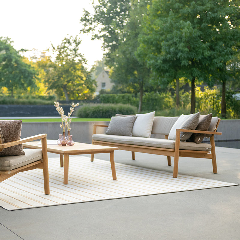 This image showcases an elegant and inviting outdoor living space, featuring a stylish seating arrangement with a natural wood-framed sofa and matching coffee table. The neutral-toned cushions and serene surrounding greenery create a harmonious and visually appealing Outdoor Furniture setting, perfect for relaxation and entertainment in a tranquil garden environment.
