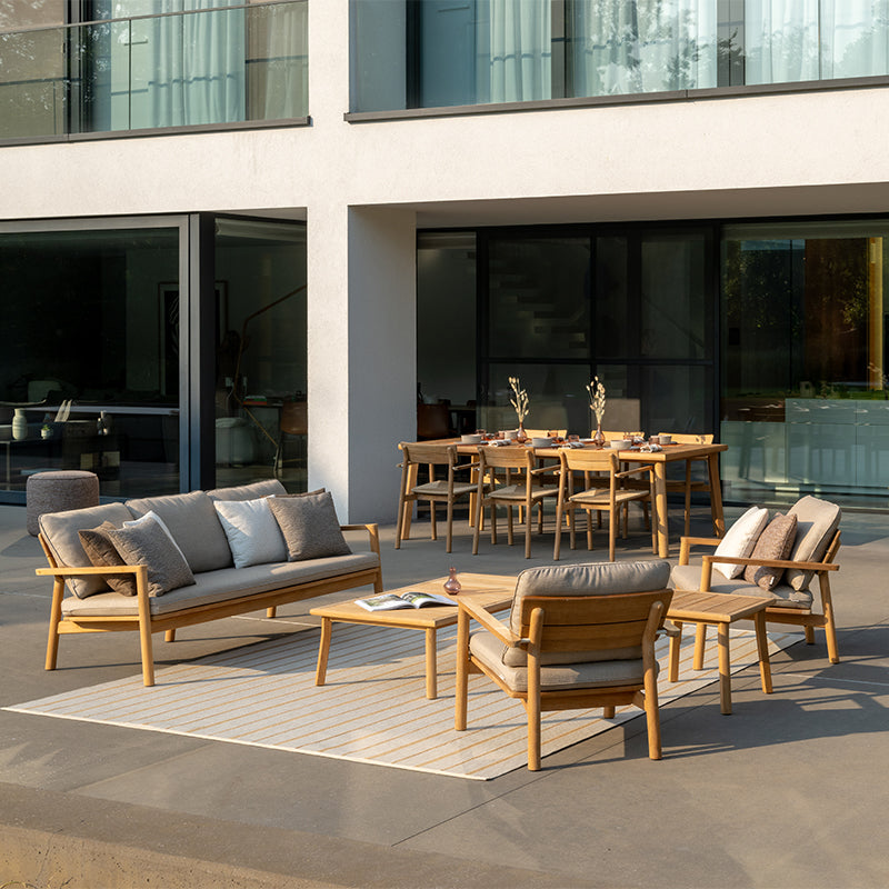 This image showcases an elegant and cohesive Outdoor Furniture arrangement, featuring a natural wood-framed sofa, armchair, and coffee table set against a backdrop of modern architecture. The neutral-toned cushions and natural finishes create a harmonious and visually appealing outdoor living space, complemented by a dining area with a wooden table and chairs. The overall design blends seamlessly with the surrounding environment, offering a comfortable and inviting space for relaxation and entertaining.