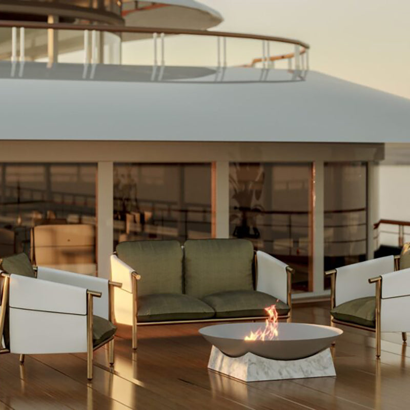 The image shows a modern, luxurious outdoor patio or lounge area. It features comfortable seating, including a green velvet loveseat and matching armchairs, arranged around a marble-topped coffee table with a built-in fire pit. The setting is surrounded by floor-to-ceiling glass walls or windows, providing a panoramic view of the outdoors. The overall atmosphere suggests an upscale, high-end design aesthetic suitable for a hotel, resort, or private residence.